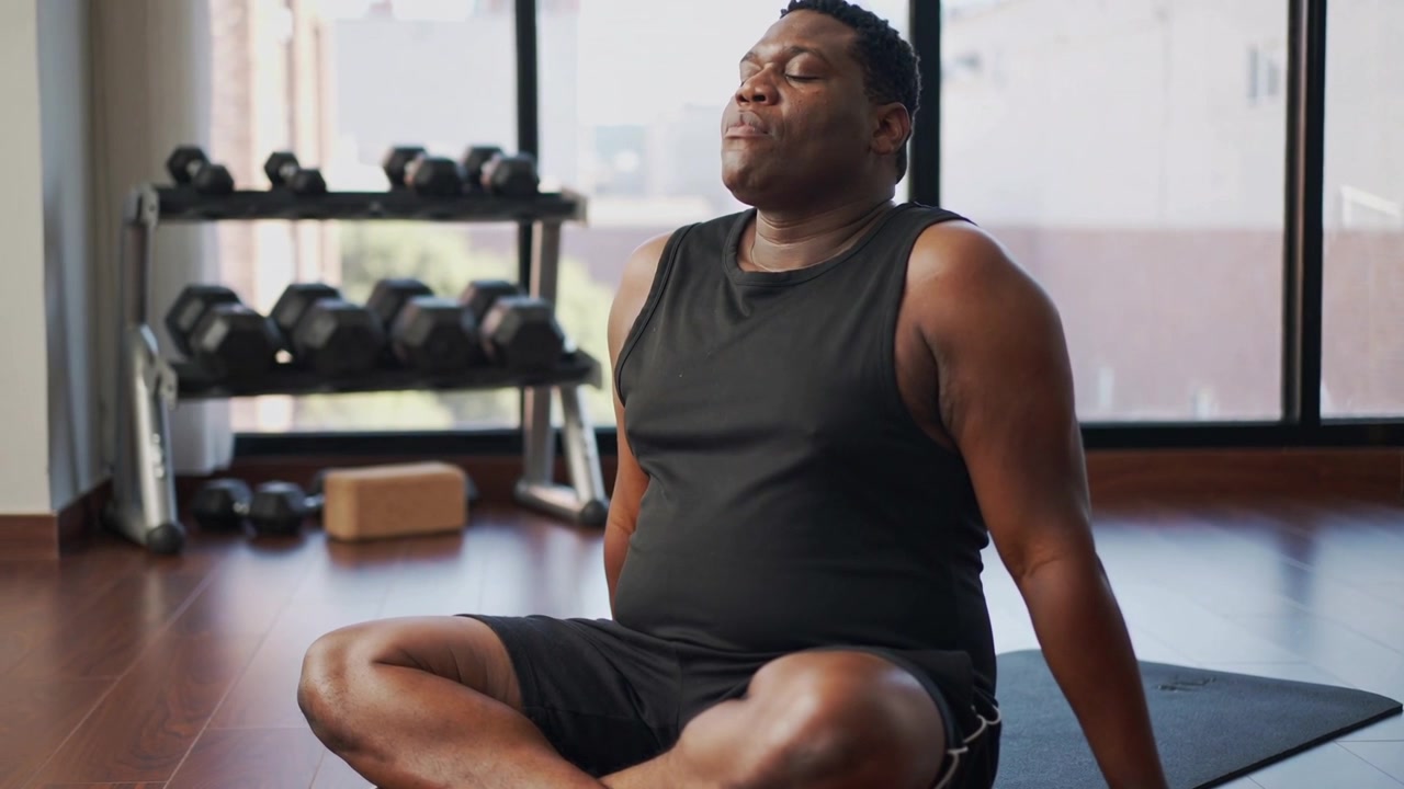 Finishing workout, man wearing black sleeveless top breathing on mat near dumbbell rack and windows