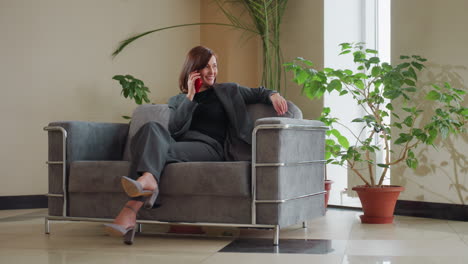 professional businesswoman in gray suit lounges on stylish couch while speaking on red smartphone in bright office filled with natural light and green potted plants, expressing calm confidence and poise