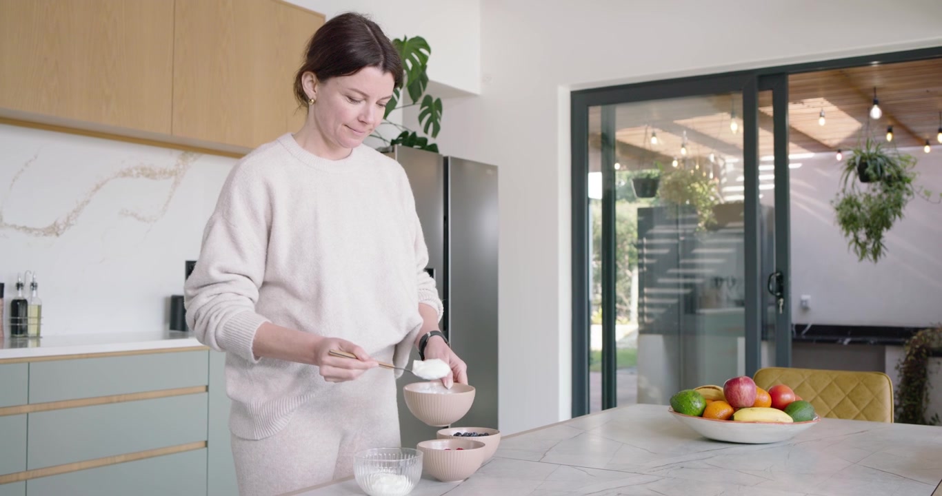 Donna adulta dopo aver ricevuto una notifica sul telefono, prepara il pasto, versa lo yogurt nelle ciotole e mescola sull'isola della cucina