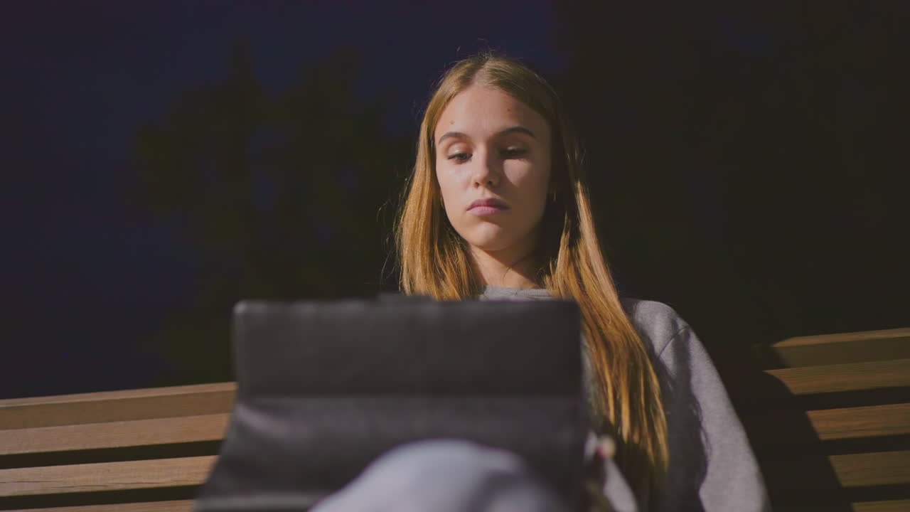 Close-up of lady sitting on park bench at night, deeply focused on tablet under soft park lighting with deep blue sky in background