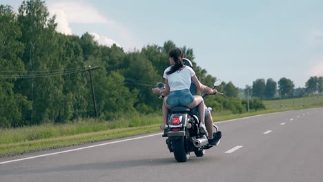 una mujer joven monta con un hombre en una motocicleta negra más allá de los árboles