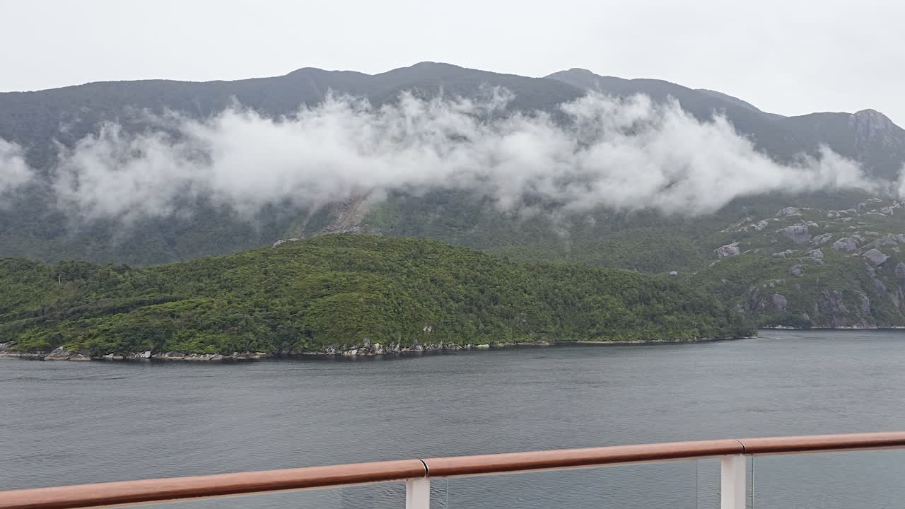 Milford Sound in Fiordland National Park, New Zealand, features cruise view of dark water fiord and steep, green mountain slopes with mist and low hanging clouds obscuring peaks