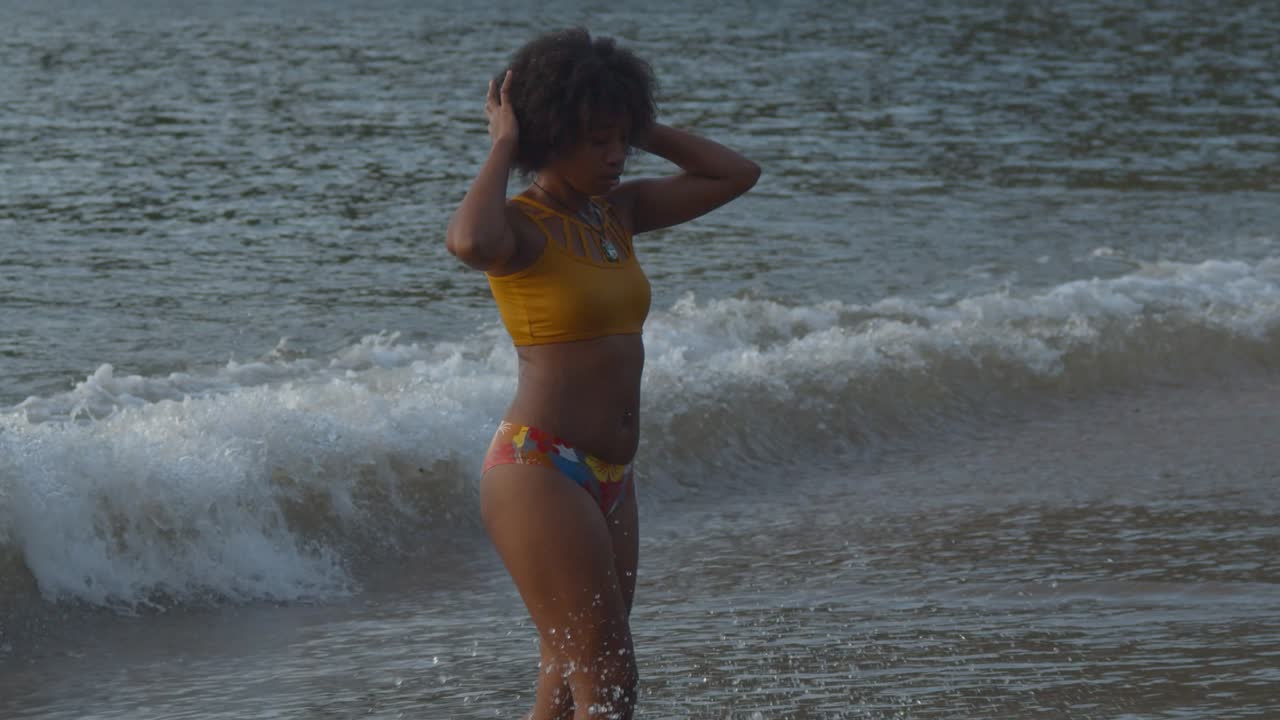 Una ragazza di colore in bikini esce dall'oceano con le onde che si infrangono sullo sfondo
