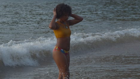 a sexy black girl in a bikini walks out of the ocean with waves crashing in the background