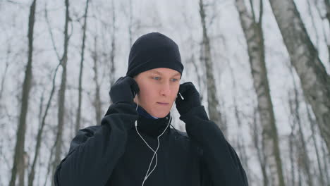 un joven que corre por la mañana sostiene auriculares en sus manos y se los inserta en los oídos antes de correr en el parque. carrera de invierno. utilice dispositivos y aplicaciones de formación modernos. escuchar música mientras entrenas mientras corres