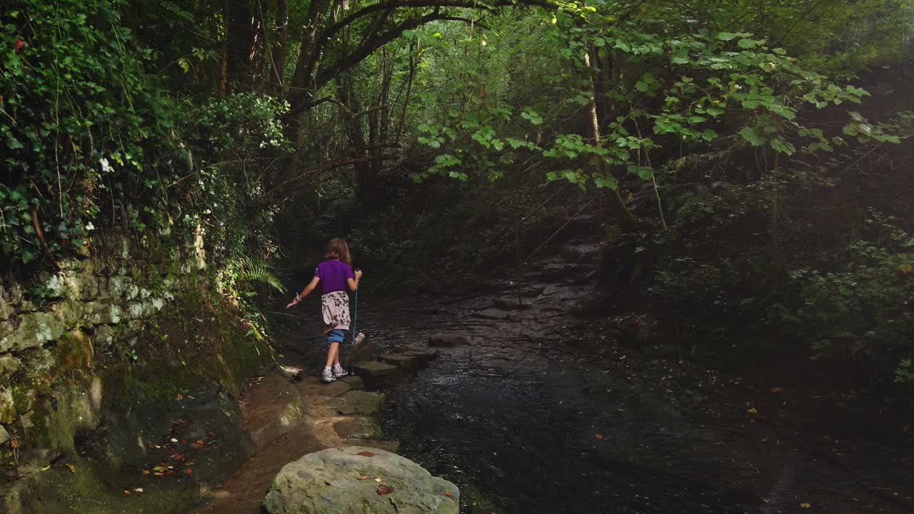 Young female explorer hiking with a pole along a scenic rocky path in a lush green forest in Rupit, Catalunya. This footage shows the concept of adventure, discovery, and active childhood