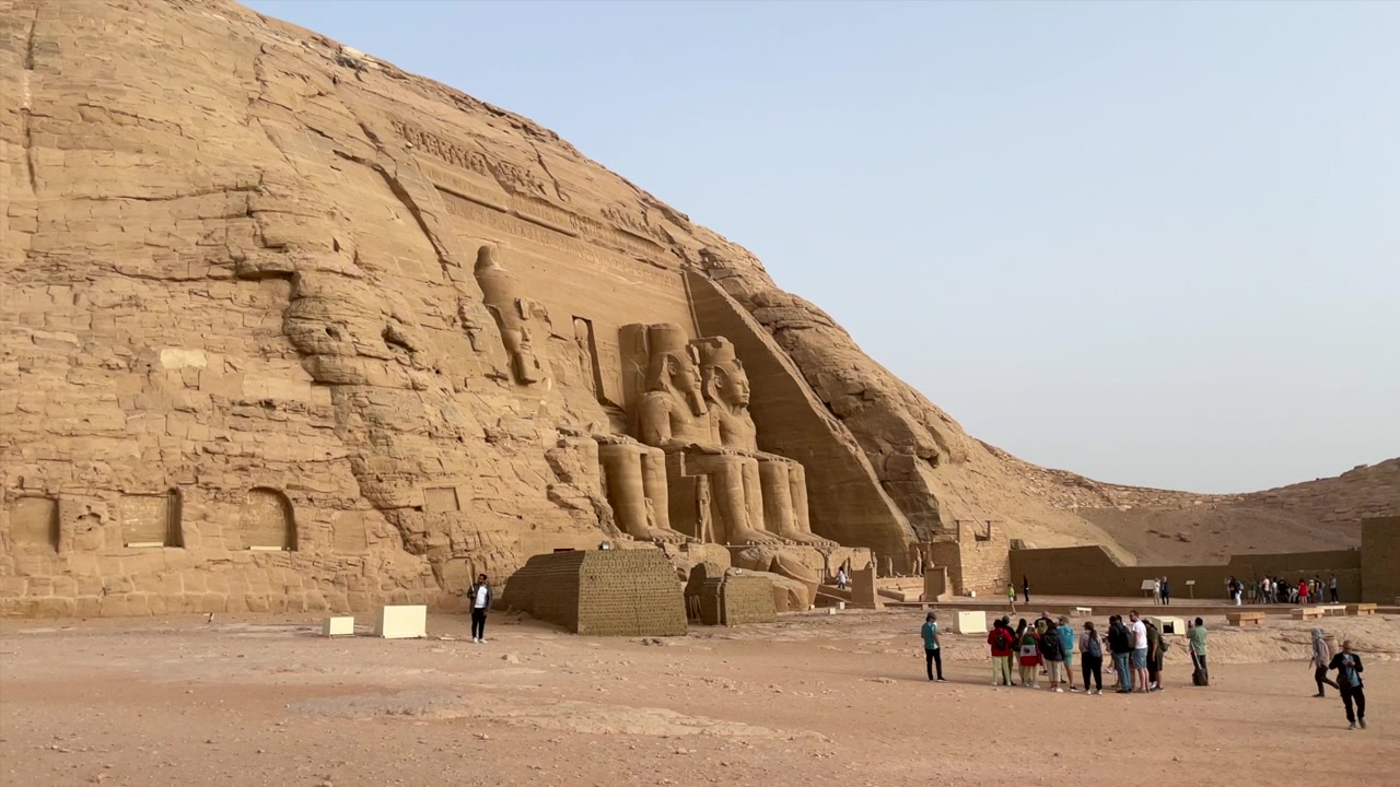 STATUA DEL FARAONE RAMSES II SUL SUO TRONO, VISTA LATERALE SINISTRA. TEMPIO IN EGITTO NEL VILLAGGIO DI ABU SIMBEL, CON TURISTI. MONUMENTI NUBIANI DELL'ANTICO EGITTO. SITO PATRIMONIO DELL'UMANITÀ UNESCO. TURISMO MONDIALE