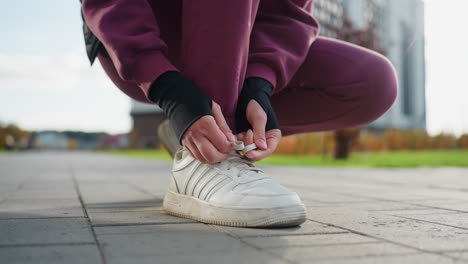 low side view of woman bending on paved walkway tying sneaker lace before run near apartment building in urban park morning light wearing maroon tracksuit and black sleeve gloves with white socks