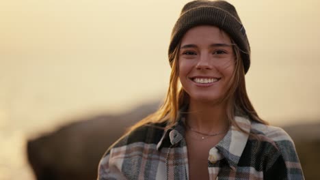 Portrait-of-a-happy-blonde-girl-in-a-black-hat-in-a-plaid-shirt-who-looks-at-the-camera-and-smiles-while-standing-on-a-rocky-shore-near-the-sea