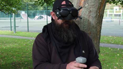 hombre barbudo con gafas fpv controlando drones en el parque con joystick de movimiento