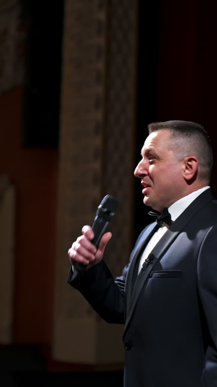 Host in elegant suit starts his speech. Speaker talking to the microphone on stage of a theater. Showman on red curtains background. Vertical video