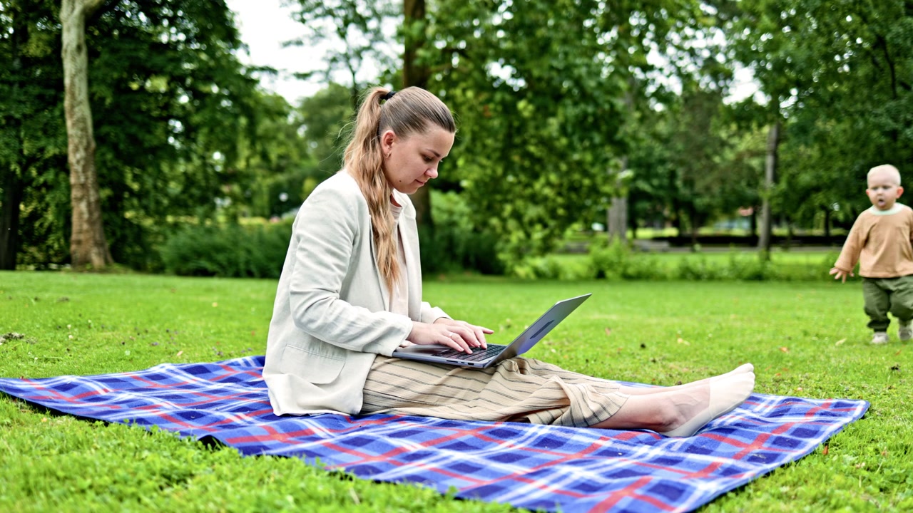 Young woman sitting in the park on green grass and working online on a laptop. Woman working remotely using a laptop. Student sitting on the lawn and typing on a laptop.