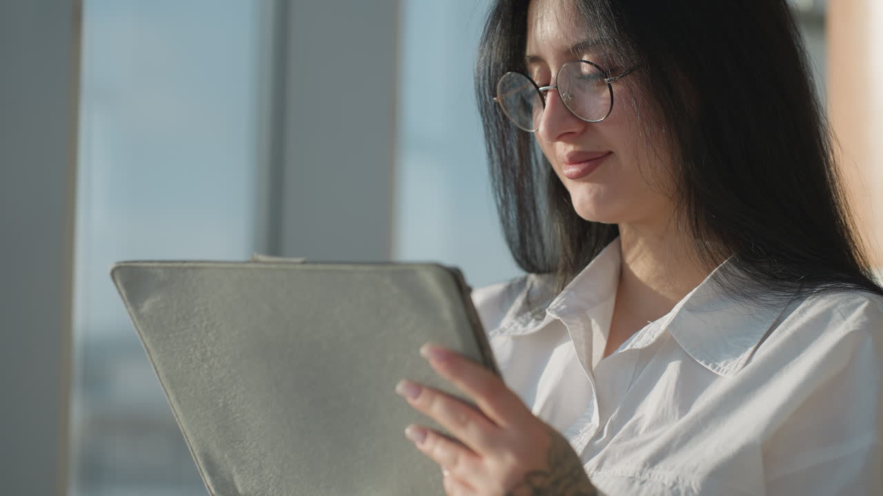 Close up beautiful smiling woman with glasses gently pushes hair back while holding tablet in brightly lit modern interior, sunlight enhancing her features and casting soft shadows