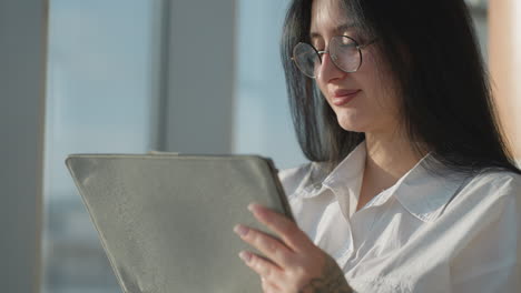 close up beautiful smiling woman with glasses gently pushes hair back while holding tablet in brightly lit modern interior, sunlight enhancing her features and casting soft shadows