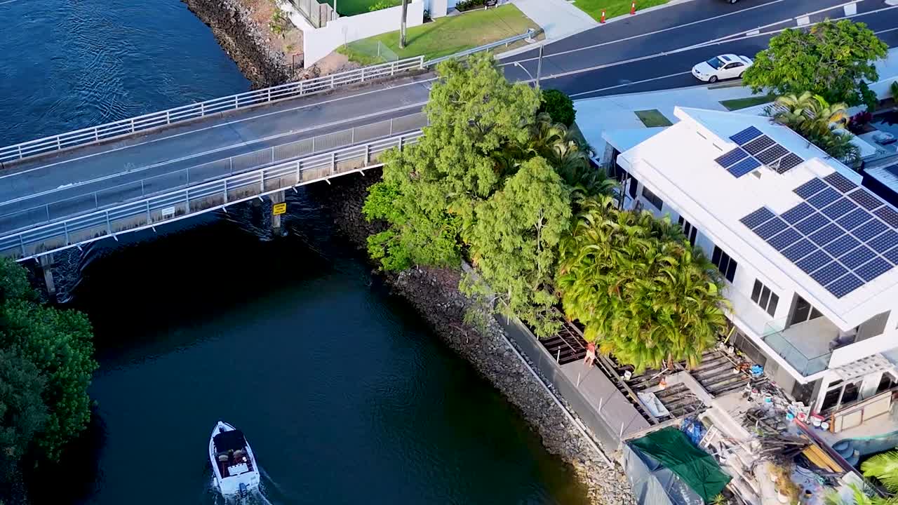 An aerial perspective of a boat passing under a bridge beside modern homes with solar panels.