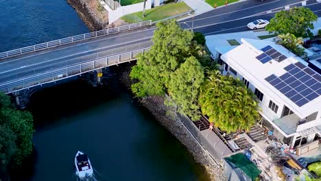 An aerial perspective of a boat passing under a bridge beside modern homes with solar panels.