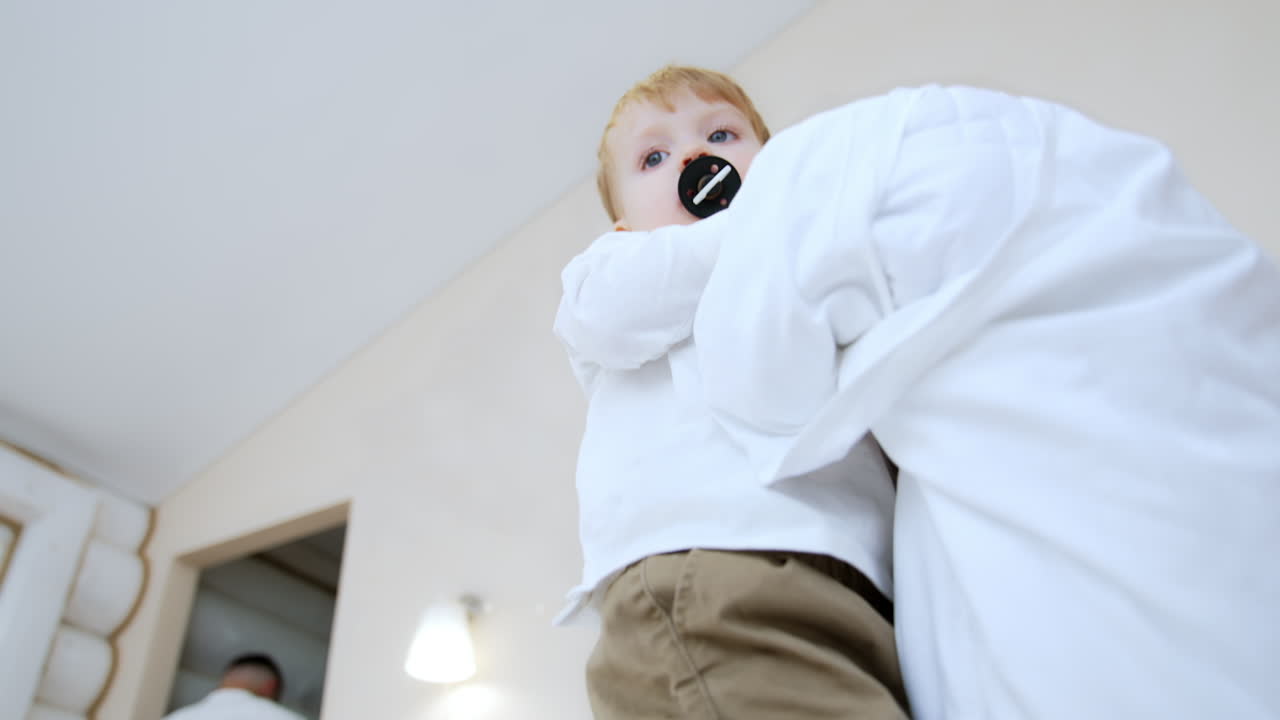 Kid with pacifier in mouth walks by the bed. Toddler boy holds a big pillow in hands. Low angle view.