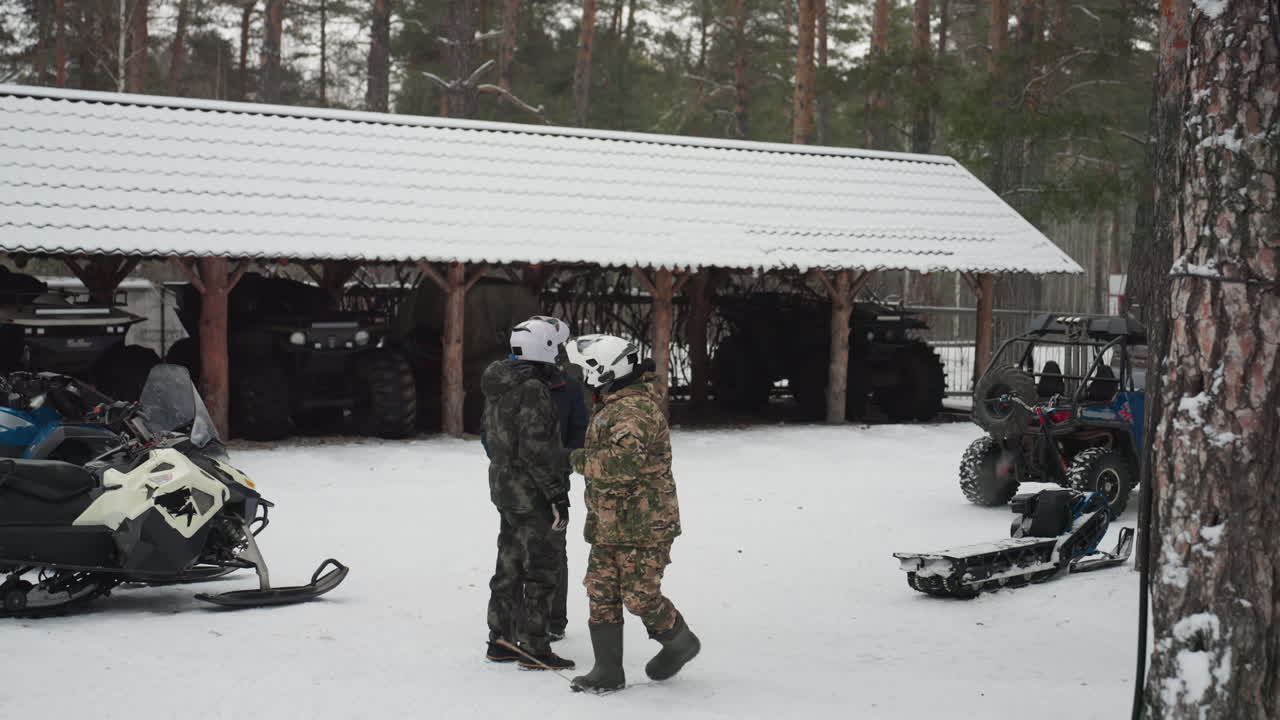 Kuljettajat lumisella tallialueella valmistellen mönkijän ja moottorikelkan varusteita. Maastokuvioitu kuljettaja neuvottelee oppaan kanssa, mekaanikko tarkastaa moottorin. Kypärät ja ajolasit näkyvissä. Mäntymetsätausta, kylmä ja pilvinen sää.