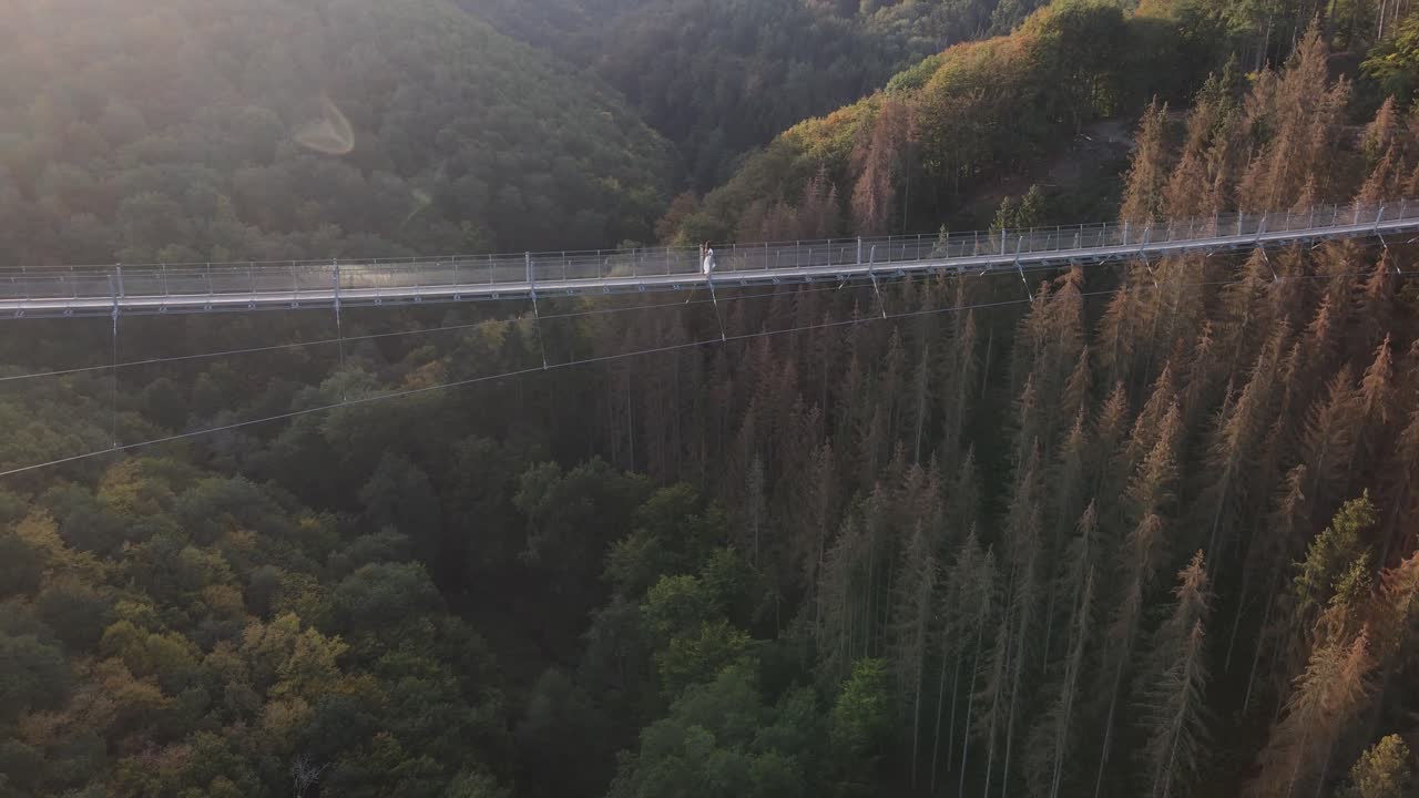 Drone footage of girl in white dress walking over suspension bridge in Germany, no color grading, flat video profile