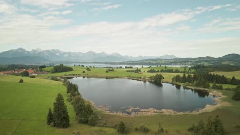 Drone nears a peaceful lake in Allgäu, Germany, capturing calm waters and the stunning Alps beyond.