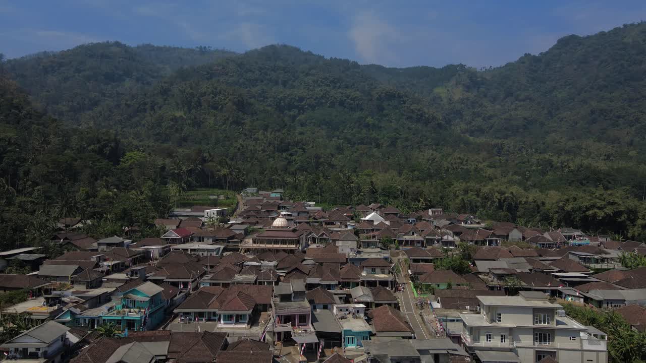 Aerial shot of Indonesian small town with clustered houses, main road, and tropical hillside background