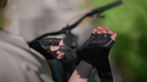 close-up of an individual standing next to bicycle, unfastening strap of biker glove with careful attention, while background showcases blurred greenery