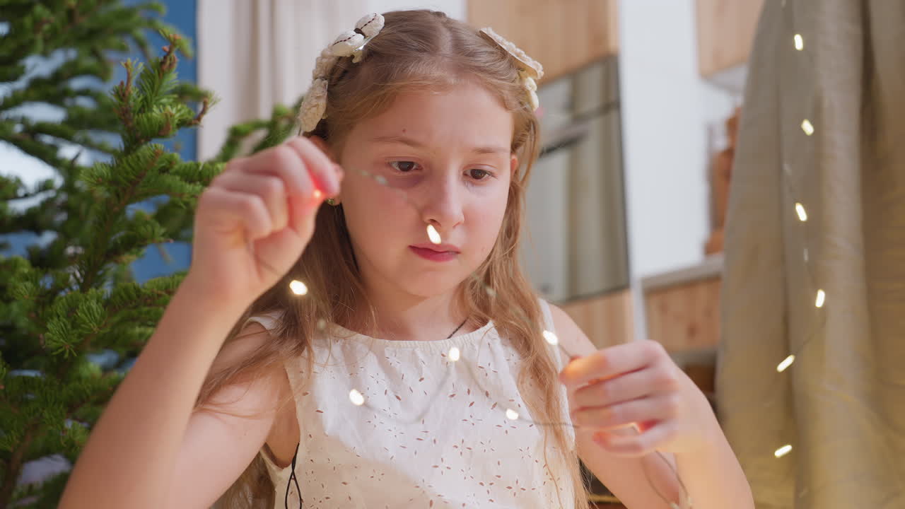 Portrait of little girl in white gown carefully twisting Christmas lights with focus on her hand movements, ready to decorate tree for Christmas holiday celebrations with family