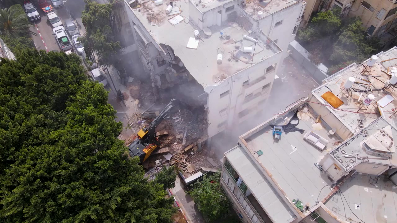 Dron girando en el aire mostrando una excavadora retirando escombros de edificios dañados por misiles.