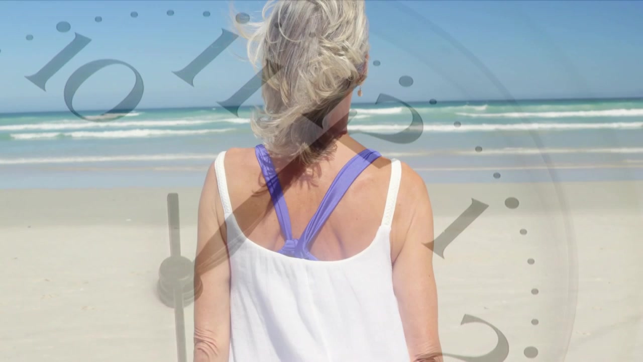 Mulher idosa em pé de frente para o oceano, com o vento levantando o cabelo, enquanto uma sobreposição de relógio marca o tempo para o bem-estar.