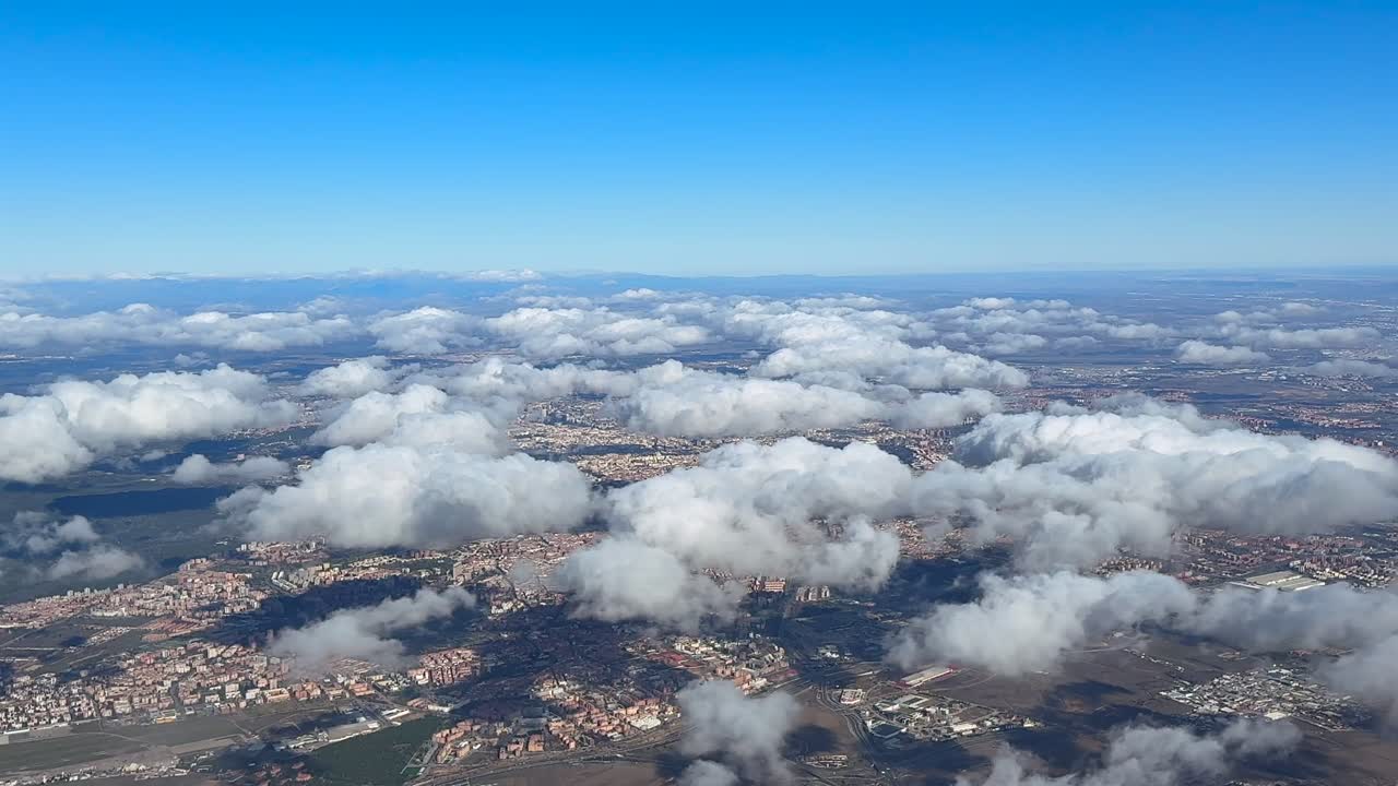 Una prospettiva elevata di Madrid scattata dalla cabina di pilotaggio, con sparsi piccoli cumuli a bassa quota sopra la città, sotto un cielo blu.