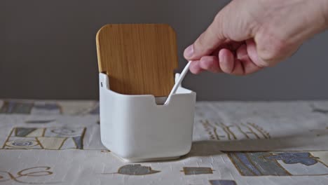 Man's hand lifts a small white ceramic spoon from sugar jar with a bamboo lid, scoops sugar, spills it back in, and replaces the spoon. Static close-up tabletop view
