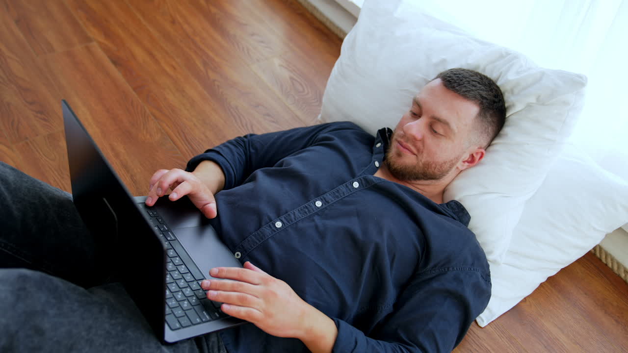 Calm bearded male lying on the floor holding legs up on the bed focused on his laptop. Top view on the man using gadget for remote work or fun at home.