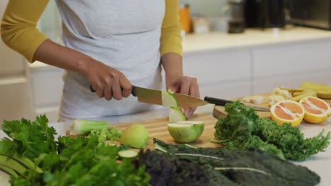 Mid-section-of-mixed-race-woman-preparing-healthy-drink,-cutting-fruit-and-vegetables-in-kitchen