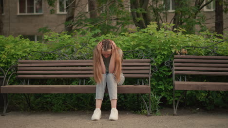 woman sits on wooden bench, resting her head in hands in a thoughtful, somber posture, surrounded by greenery and an urban building in background. her long hair flows down as she adjusts her head