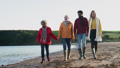 Ein-älteres-Paar-Hält-Sich-An-Den-Händen,-Während-Es-Mit-Seinem-Erwachsenen-Nachwuchs-Im-Winterlichen-Strandurlaub-An-Der-Küste-Entlang-Spaziert