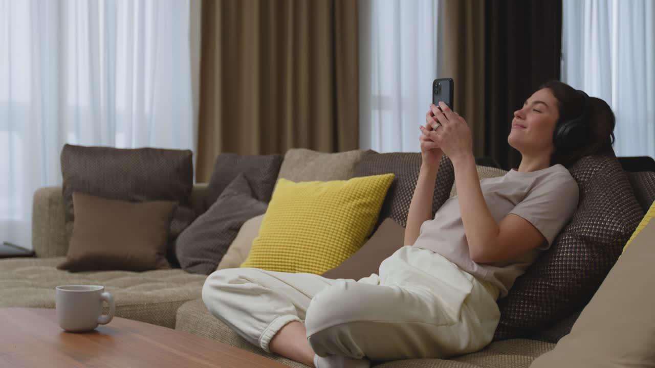 집에서 소파에 앉아 휴대전화 음악 앱을 사용하여 헤드폰으로 좋아하는 음악을 듣는 현대적인 젊은 여성