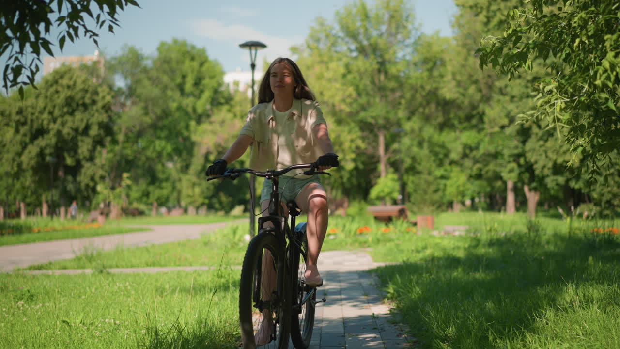 젊은 여성은 초록색으로 가득 찬 경치 좋은 공원의 포장된 길을 자신감 있게 자전거를 타고, 빛은 나무를 통해 필터링, 길에 얼룩진 그림자를 던집니다.