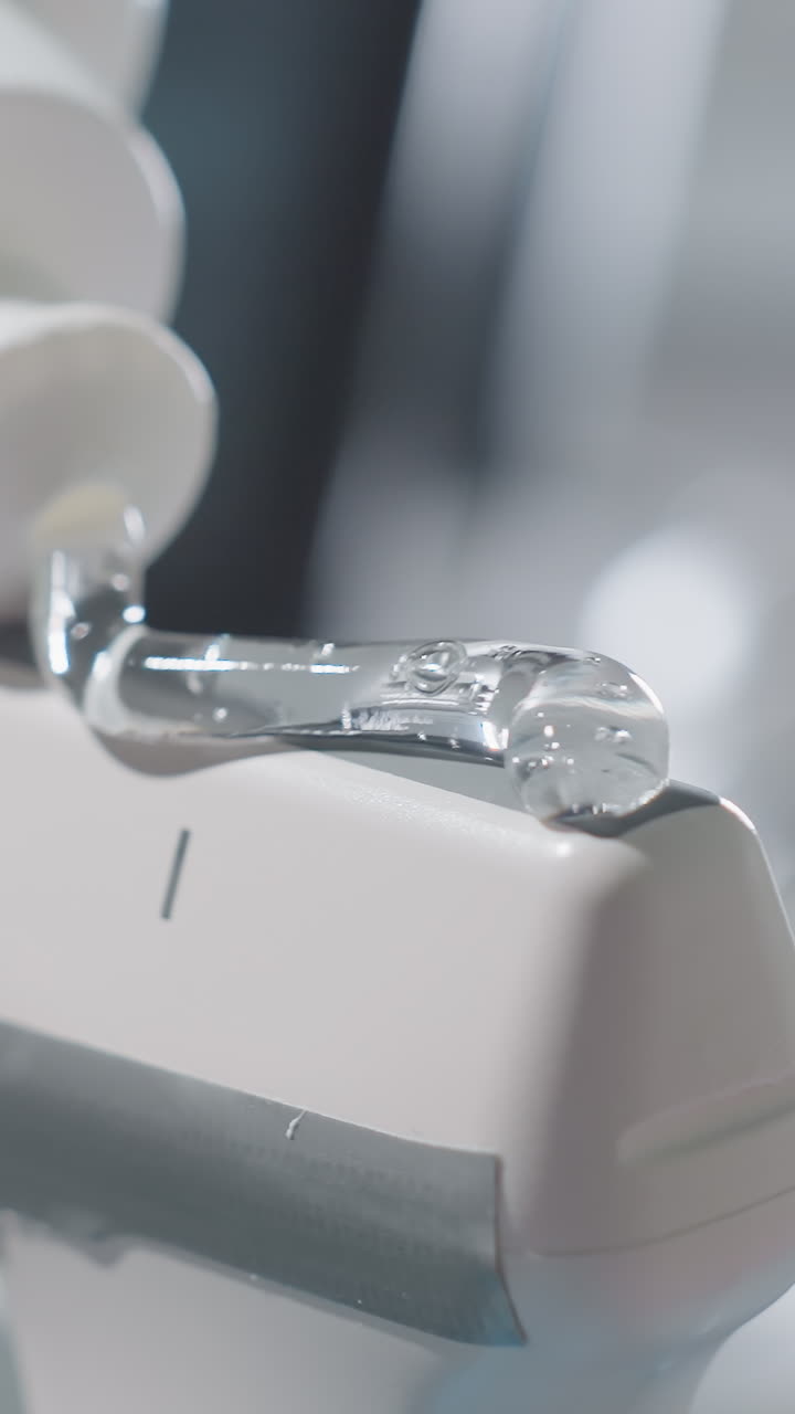 초음파 탐촉자에 테크니션이 디스펜서를 사용하여 젤을 바르는 클로즈업 장면입니다. 투명한 전도성 젤 방울이 광택 있는 변환기 표면에 퍼져 있고, 클리닉 모니터 보케와 살균된 공간에 초점이 맞춰져 있습니다.