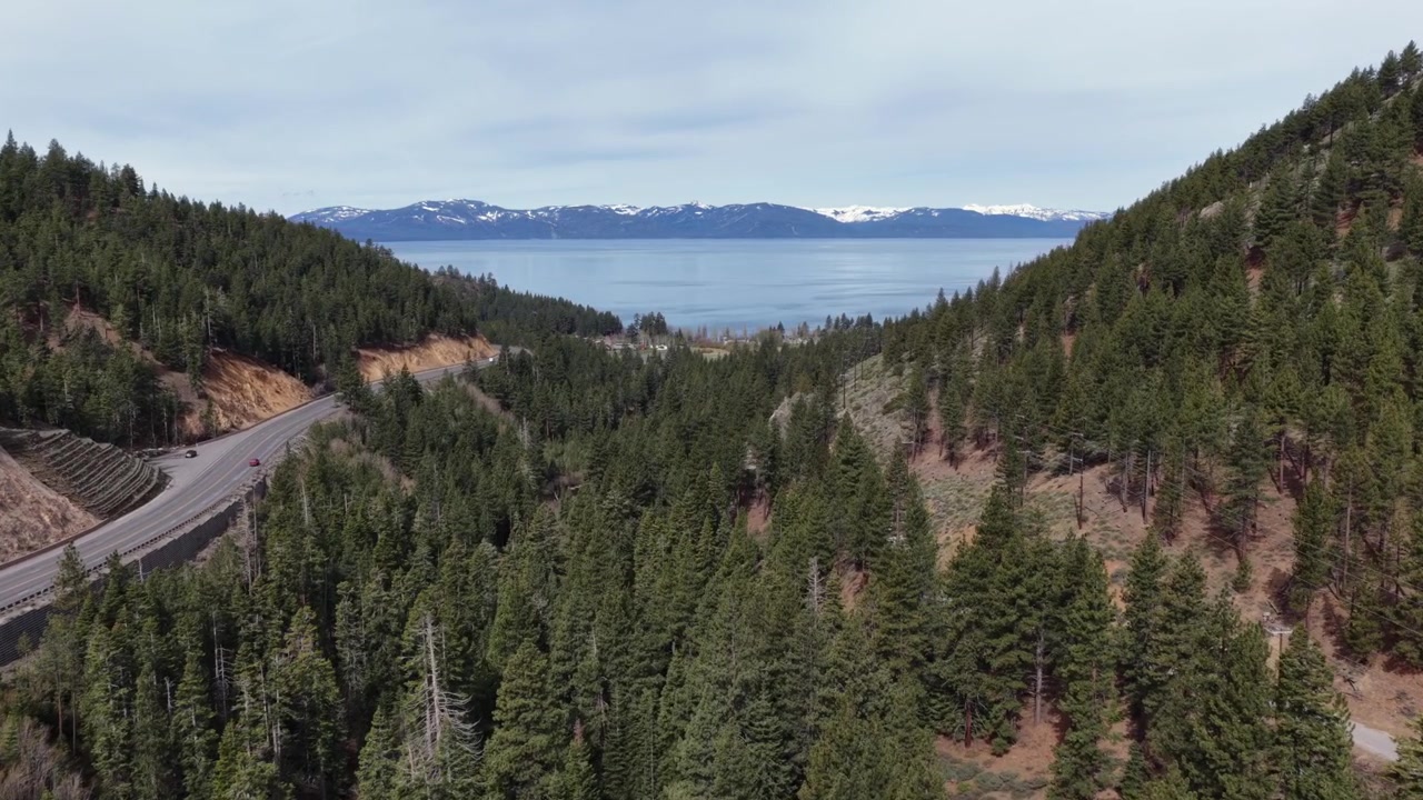 Lake Tahoes betagende panoramaudsigt med dybblå alpesø, sneklædte Sierra Nevada-bjerge, tæt fyrreskov og snoet bjergmotorvej langs en stenet bjergside. Bred luftoptagelse.