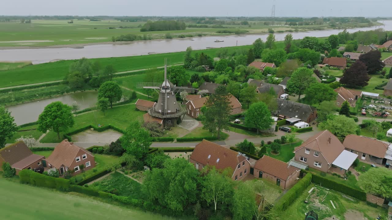 Aerial shot ascending over a peaceful countryside with windmill and red-roof houses. Scene captured during daylight under overcast skies.