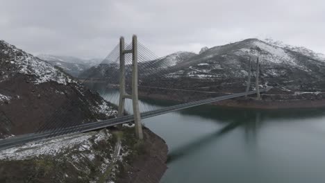 Puente-Camino-Cruzando-Cordillera-Sobre-El-Río-Paisaje-Invernal-Embalse-De-Barrios-De-Luna-En-León,-España-Vista-Aérea-De-órbita-De-Drones