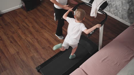 top view of young boy gripping treadmill handles while engaged in intense indoor workout session, demonstrating focus and energy as adult supervises activity in cozy home setting