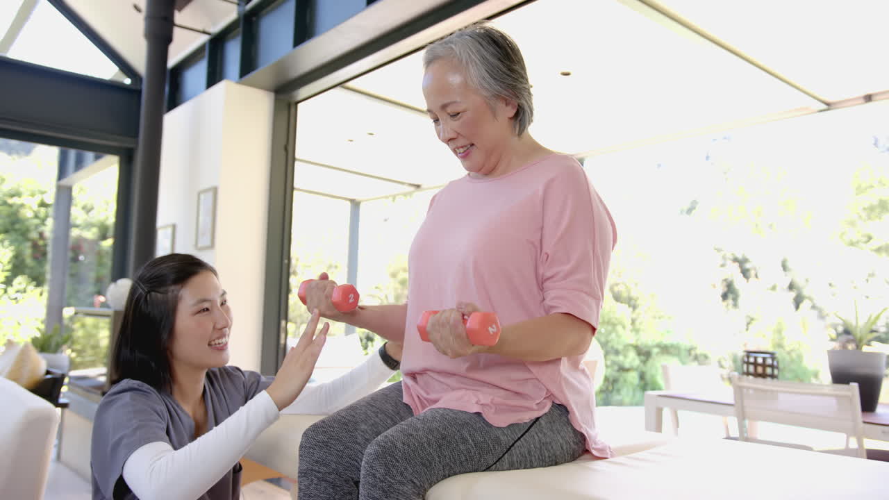 Assisting senior asian woman with dumbbells, physiotherapist encouraging during home workout