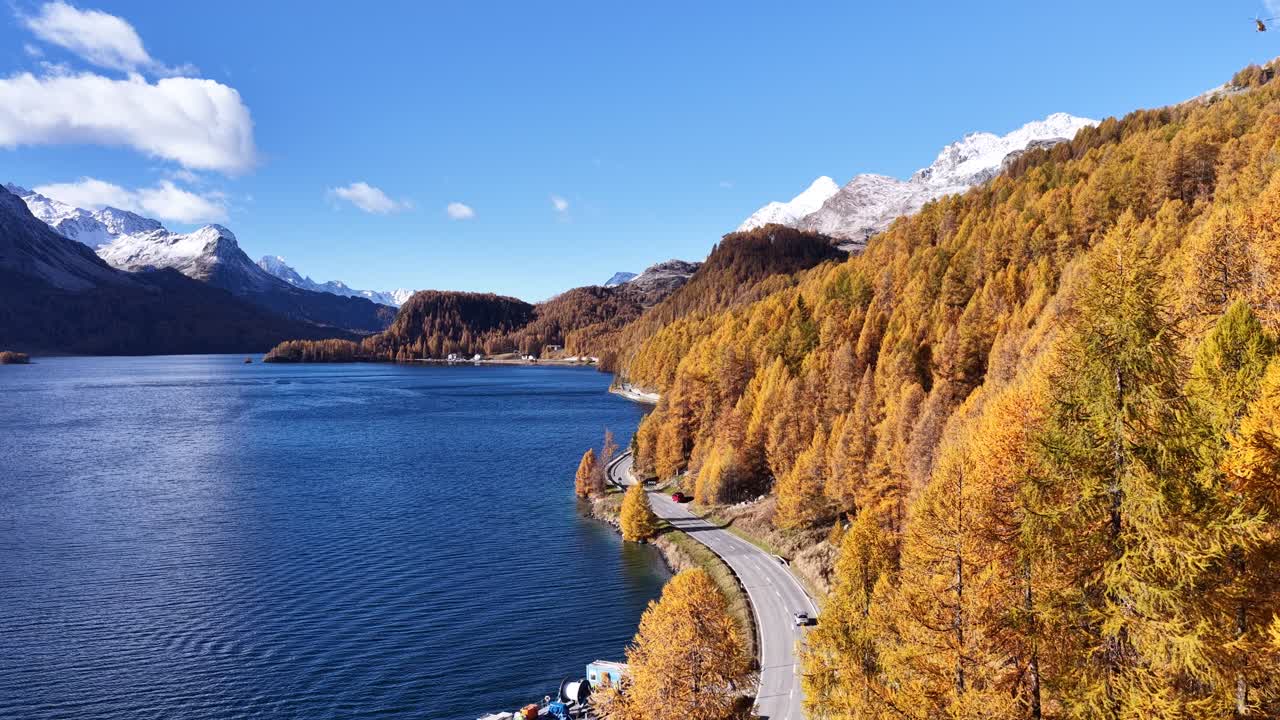 Aerial view of Alpine lake with golden larch trees, mountain road with cars, and snow-capped peaks