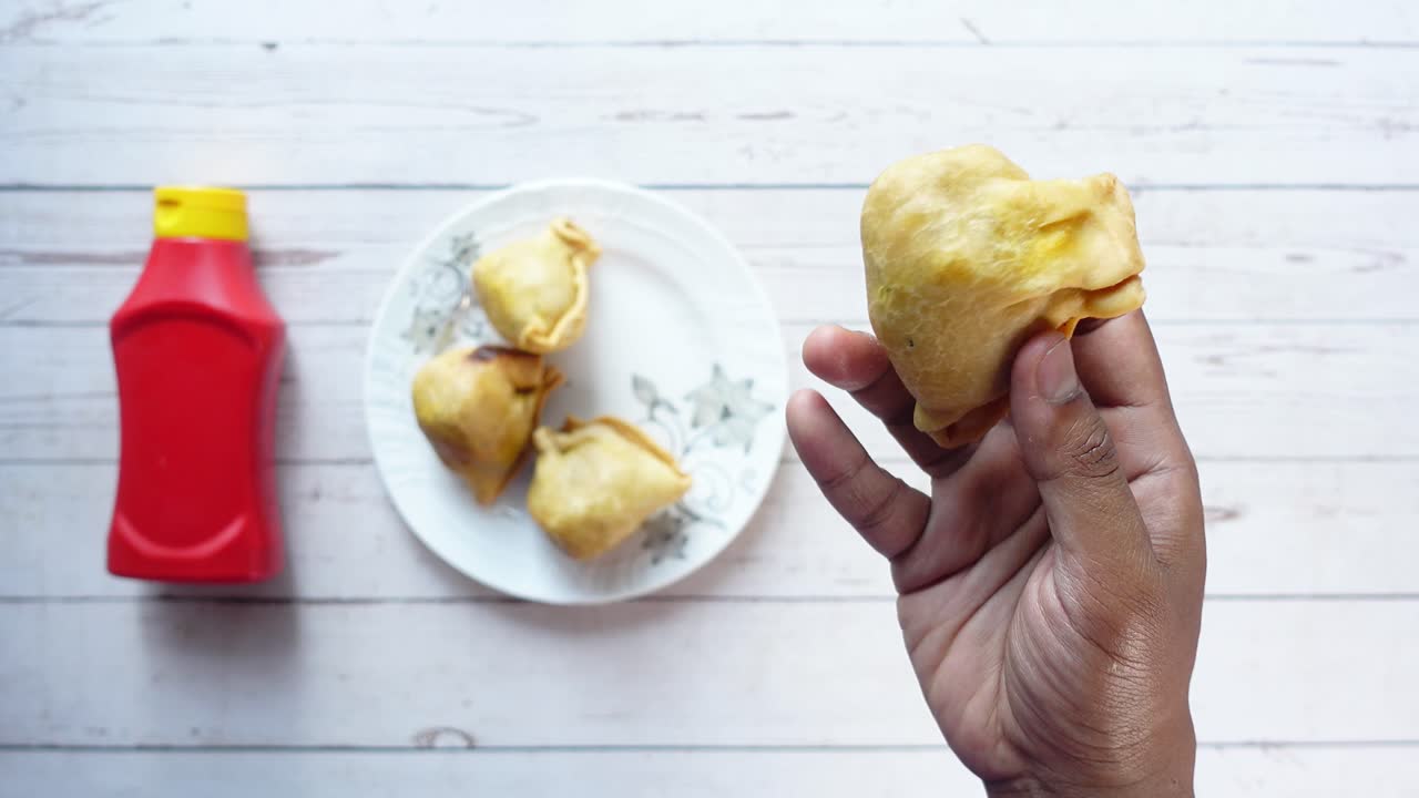 una mano sosteniendo una samosa con un plato de samosas y ketchup en el fondo