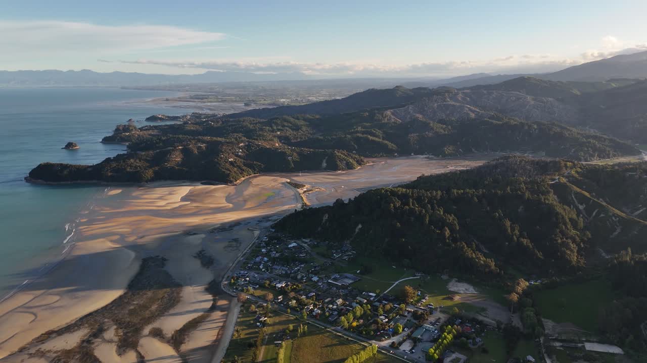 Yeni Zelanda, Güney Adası'nda gün batımında Marahau kasabası ve Abel Tasman kıyı şeridinin panoramik hava görüntüsü; altın rengi gelgit kumulları, turkuaz okyanus, dalgalı tepeler ve sıcak akşam ışıkları altındaki köy ile.