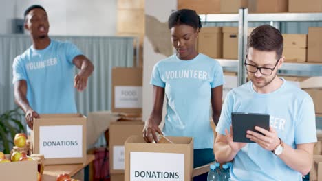 multiethnic group of volunteers packing boxes with food and clothes and typing on tablet in charity warehouse