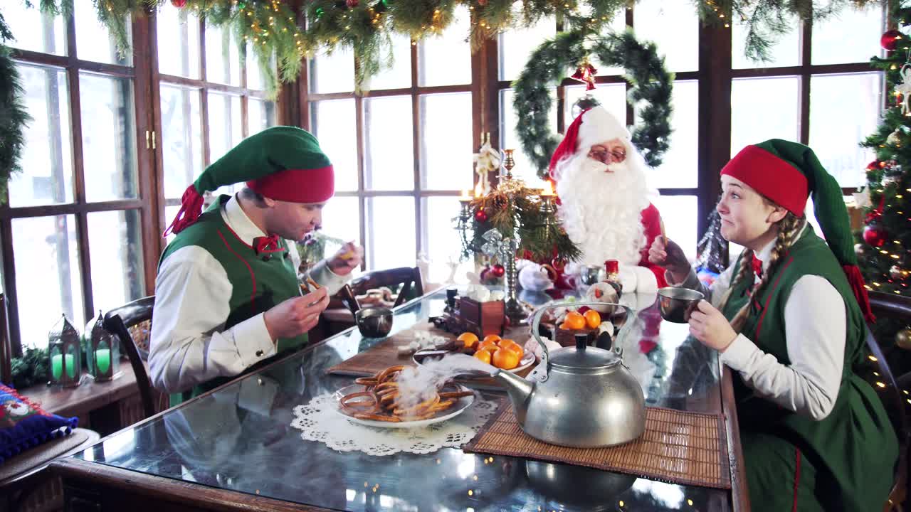 Hungry elves eating with Santa at festive table. Two elves drinking tea with Santa in a room with Christmas decorations