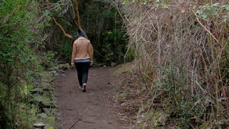young winter dressed brunette walking through a path surrounded by green forest getting away from the camera
