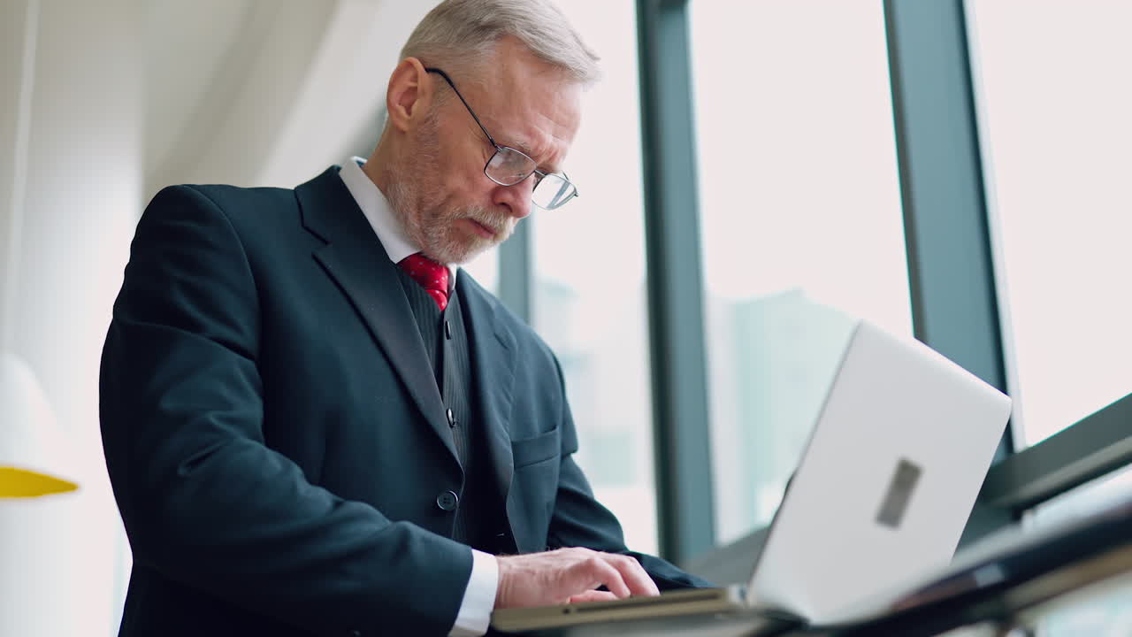 Senior serious businessman standing near window with panoramic city view. Laptop in hands, man thinking while checking e-mails standing near window. Video from the side.